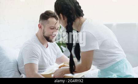 brunette woman with braids bringing breakfast tray to boyfriend in bed Stock Photo