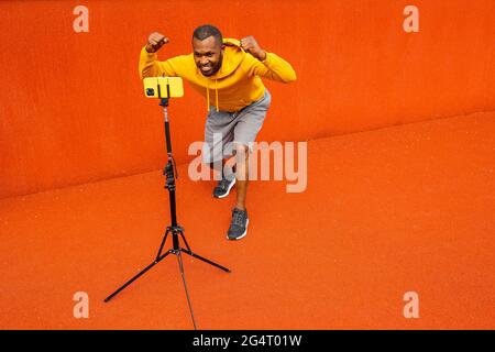 Joyful African-American sportsman athletic coach in trendy sportswear , give online lesson on mobile phone on tripod, smiling and making yes gesture Stock Photo