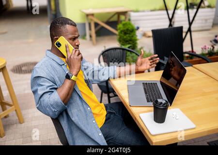Pensive afro american businessman in stylish clothing talking on mobile phone and using laptop outdoors cafe, drinking take away coffee discussing Stock Photo