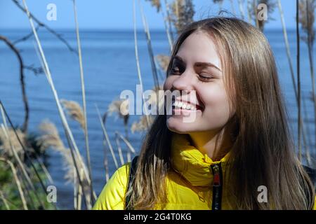Portrait of happy smiling young woman with her eyes closed enjoys the sunshine and fresh air. She stands in against lake on warm spring day in yellow Stock Photo