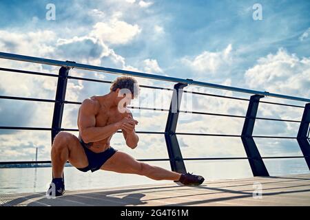Young muscular tattooed man doing exercises, stretching early in the morning on pier by the sea Stock Photo