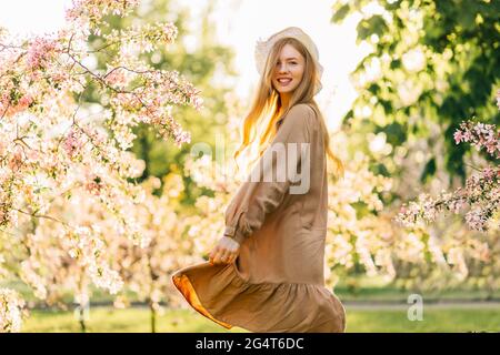Beautiful young woman in a summer hat and a delicate beige dress dancing in blooming gardens, happy woman under blooming trees in spring Stock Photo