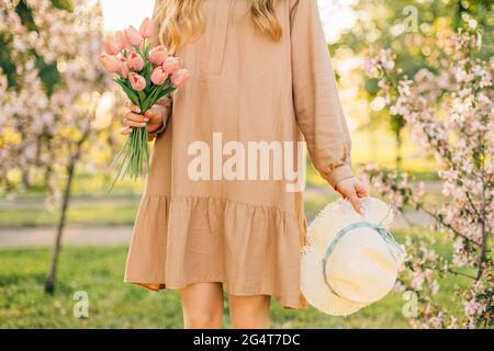 Woman with a bouquet of tulips and a hat in her hands, in a blooming spring garden, walking in a park with blooming trees Stock Photo