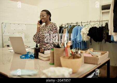 Smiling African female fashion designer talking on the phone in her workshop Stock Photo