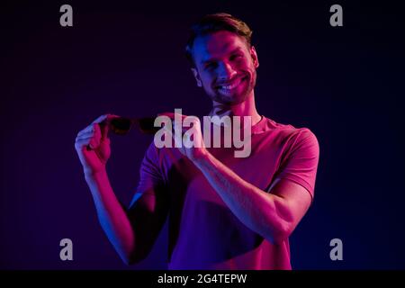 Portrait of attractive cheerful successful guy holding specs in hands isolated over dark neon purple color background Stock Photo