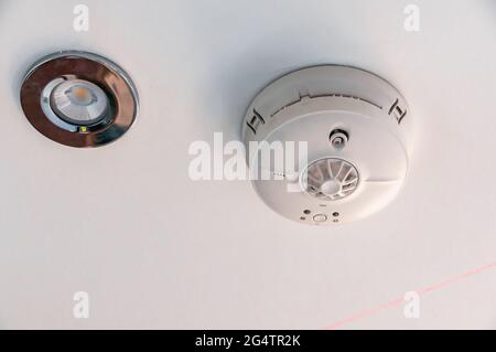 A smoke detector alarm is mounted on a white kitchen ceiling next to an inset LED downlighter Stock Photo