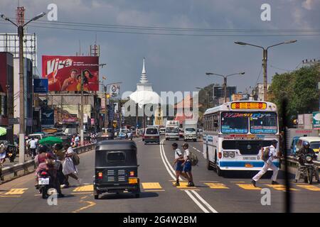 View down a main road with traffic and a pedestrian crossing with people on, near Columbo, with street lights, hoardings and temple dome Stock Photo