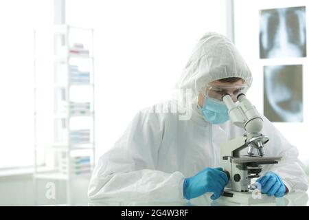 Doctor in a protective suit with gloves adjusts protective glasses near a microscope in a medical laboratory Stock Photo