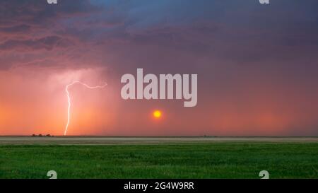 Lightning bolt strikes at sunset with the sun seen heavily filtered by the rain falling in the distance. Dark storm clouds can be seen overhead. Stock Photo