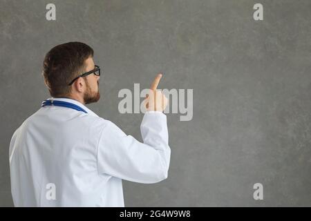 Focused male doctor as if reading something on a gray background pointing a finger at it. Stock Photo