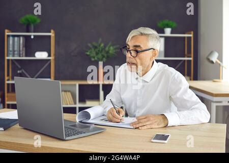Busy senior male worker sitting at laptop making notes while working in modern coworking center. Stock Photo