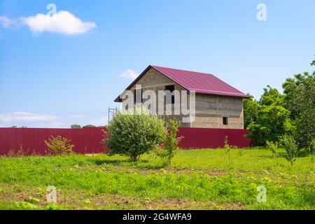 Private house under construction behind a fence. In the foreground is a green garden. Countryside and construction. Stock Photo