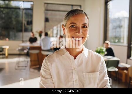 Mature woman standing in co-working space with people discussing in background. Senior businesswoman standing in office and colleagues working at back Stock Photo