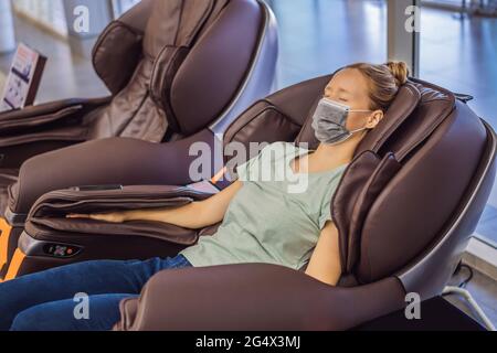 Beautiful young woman wearing a medical mask due to the coronavirus COVID 19 relaxing on the massage chair in airport or in the mall Stock Photo