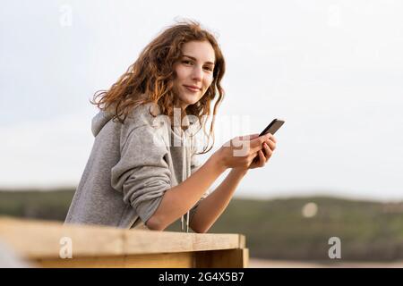 Redhead woman holding mobile phone while leaning on railing by sky Stock Photo