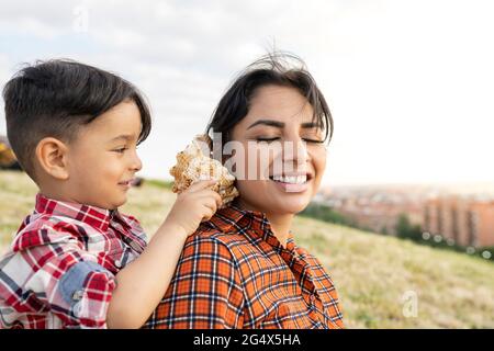 Smiling woman listening to conch shell with boy behind on hill Stock Photo