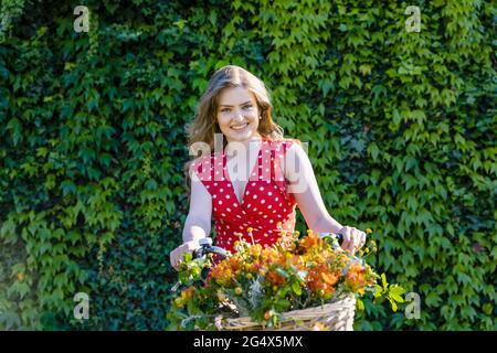 Smiling beautiful woman sitting on bicycle with basket with freesia flowers in front of ivy hedge Stock Photo