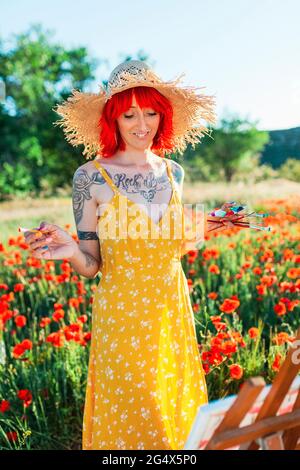 Smiling woman in sundress holding paintbrushes while looking at canvas on field Stock Photo