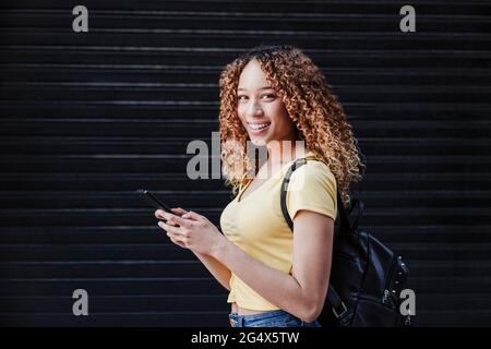 Young woman with backpack holding smart phone in front of shutter Stock Photo