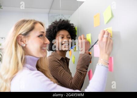 Smiling female colleagues writing on adhesive notes together while discussing in office Stock Photo