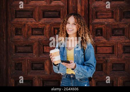 Young woman with reusable cup holding smart phone while standing in front of wooden door Stock Photo