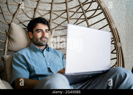 Male freelancer using laptop while sitting on chair swing in cafe Stock Photo