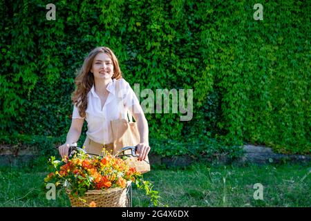 Happy beautiful woman sitting on bicycle with basket of freesia flowers in front of green ivy hedge Stock Photo