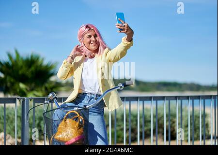 Smiling young woman taking selfie through smart phone while sitting on bicycle Stock Photo