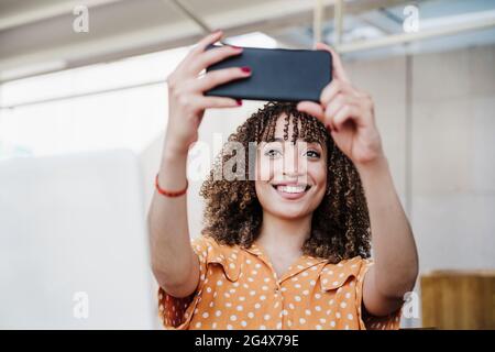 Smiling young woman taking selfie through smart phone in cafe Stock Photo