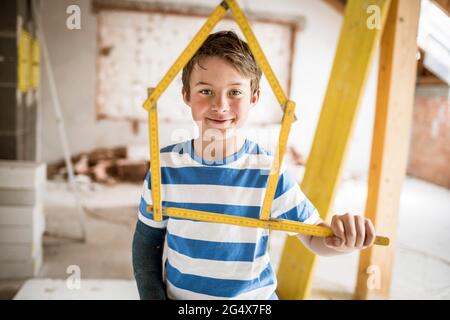 Smiling boy holding tape measure of house shape at attic Stock Photo