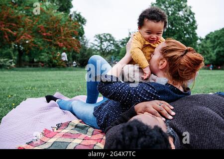 Mother kissing son while leaning on man resting on picnic blanket at park Stock Photo