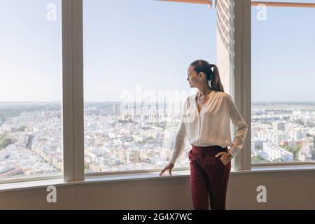 Mature businesswoman looking through window while standing with hand in pocket at window Stock Photo