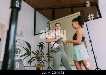 Girl showing thumbs up while standing near female friend vlogging at home Stock Photo