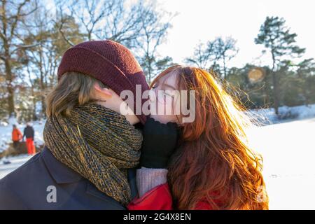 Redhead woman pulling knit hat while kissing boyfriend on sunny day Stock Photo