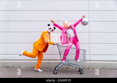 Man and woman wearing vibrant suits and animal masksÂ messing around with shopping cart and disco ball Stock Photo