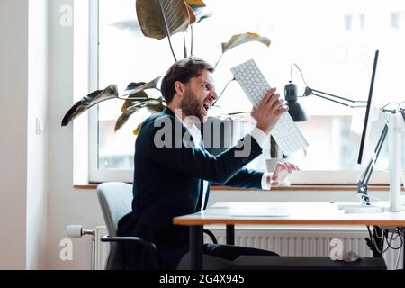Angry businessman holding computer keyboard while sitting by desk Stock Photo