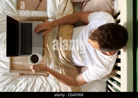 Man sitting with coffee cup and laptop on table in bed Stock Photo