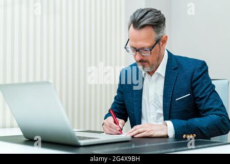 Businessman with laptop writing in notepad while working at office Stock Photo