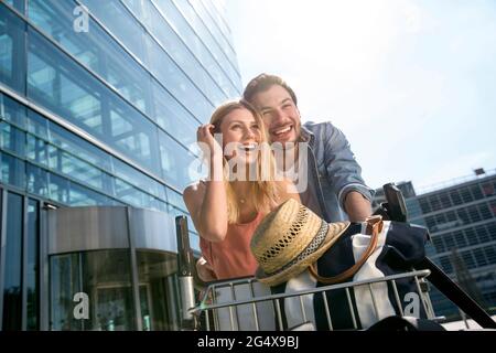 Cheerful young couple with luggage trolley standing outside airport terminal Stock Photo