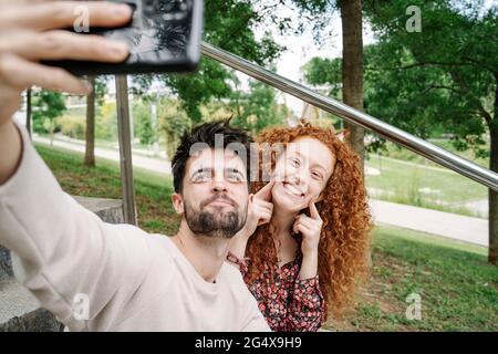 Young couple making cute faces while talking selfie in public park Stock Photo