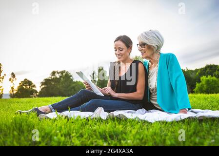 Smiling granddaughter using digital tablet while sitting by grandmother on picnic blanket Stock Photo