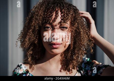 Young curly haired woman standing with hand in hair during sunny day Stock Photo