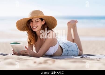 Smiling beautiful woman with book lying on towel at beach during sunny day Stock Photo