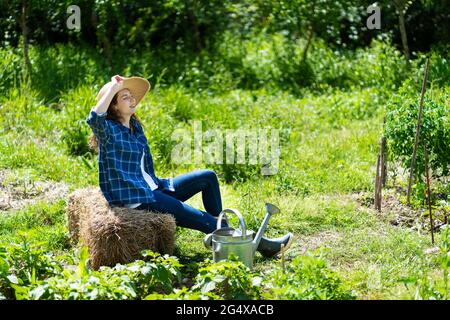 Woman sitting on stack of hay in permaculture garden during sunny day Stock Photo