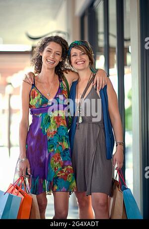 Smiling female friends carrying shopping bags while standing with hands on shoulders Stock Photo