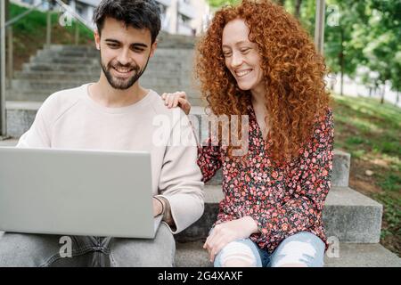 Smiling young couple using laptop while sitting on steps in public park Stock Photo