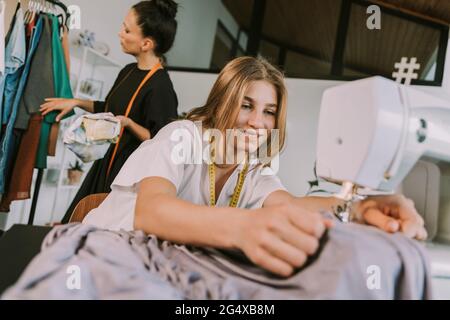 Smiling teenage fashion designer sewing cloths with mature woman in background working at workshop Stock Photo