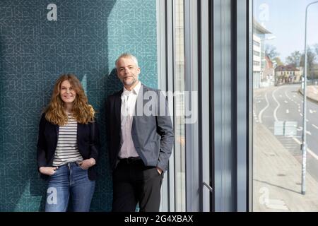 Smiling male and female professionals with hands in pockets standing side by side at office Stock Photo