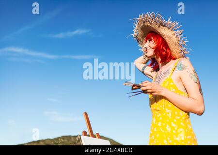 Tattooed female artist with paintbrushes looking away while holding sun hat Stock Photo