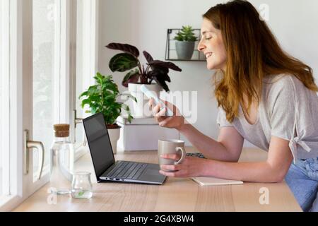 Smiling woman using smart phone over laptop while leaning on desk at home office Stock Photo